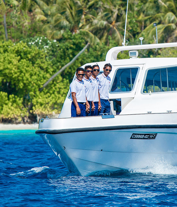 Speedboat transfer Maldives- Four uniformed crew members on a white resort speedboat speeding across turquoise waters towards a lush green Maldivian island.